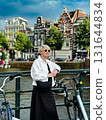Fashionable woman poses by scenic canal, with bicycles and blooming flowers nearby, showcasing a relaxed urban atmosphere and vibrant lifestyle 131644834