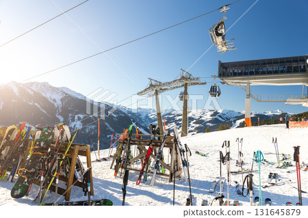 Sunny winter day on alpine slope with skis and snowboards resting near gondola station. Bright sunlight, blue sky, and mountain panorama create atmosphere of adventure, freedom, and relaxation 131645879