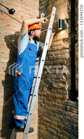 Engineer using ladder to repair lighting system on brick wall Engineer using ladder to repair lighting system on brick wall 131648356