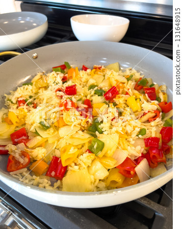 A white skillet on a gas stove holds scrambled eggs mixed with colorful red and yellow bell peppers. Kitchen bowls are placed in the background. A white skillet on a gas stove holds scrambled eggs mixed with colorful red and yellow bell peppers. Kitchen bowls are placed in the background. 131648915
