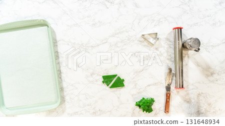 Overhead view of Green Gingerbread Cookies dough, partially rolled out, next to tree-shaped cutouts on a baking sheet and baking tools. Overhead view of Green Gingerbread Cookies dough, partially rolled out, next to tree-shaped cutouts on a baking sheet and baking tools. 131648934