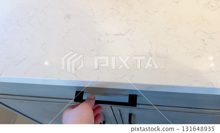 A hand pulls open a kitchen drawer that sits under a pale marble countertop. The handle is black and rectangular, contrasting with the soft tones of the cabinetry and counter. A hand pulls open a kitchen drawer that sits under a pale marble countertop. The handle is black and rectangular, contrasting with the soft tones of the cabinetry and counter. 131648935