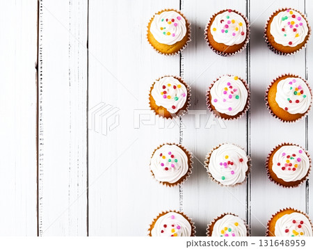 A top-down view of neatly arranged cupcakes, frosted with white icing and decorated with colorful sprinkles. The cupcakes are placed in rows on a white wooden background, creating a simple and 131648959