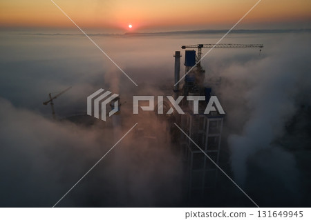 Aerial view of cement factory with high concrete plant structure and tower crane at industrial manufacturing site on foggy evening. Production and global industry concept. Aerial view of cement factory with high concrete plant structure and tower crane at industrial manufacturing site on foggy evening. Production and global industry concept. 131649945