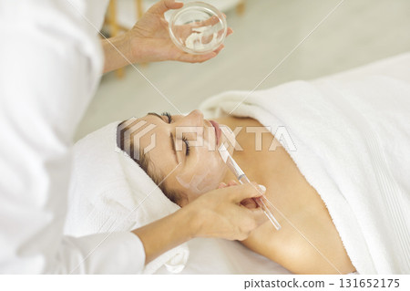 Professional cosmetologist applying moisturizing mask with brush to a young woman in spa salon. Professional cosmetologist applying moisturizing mask with brush to a young woman in spa salon. 131652175