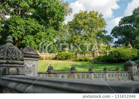 Ornate stone railings frame a grassy area surrounded by lush trees in Central Park, New York. The historic architecture contrasts beautifully with the vibrant summer landscape. Ornate stone railings frame a grassy area surrounded by lush trees in Central Park, New York. The historic architecture contrasts beautifully with the vibrant summer landscape. 131652846