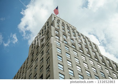 Low-angle view of a beige office building with many square windows and an American flag waving on top. The bright sky with scattered clouds highlights the sharp architectural lines of the structure. 131652863