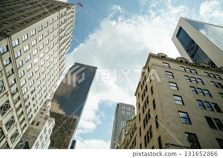 Upward view of modern and historic buildings in Manhattan with an American flag waving high above. The glass and stone facades contrast beautifully under a partly sunny sky. Upward view of modern and historic buildings in Manhattan with an American flag waving high above. The glass and stone facades contrast beautifully under a partly sunny sky. 131652866