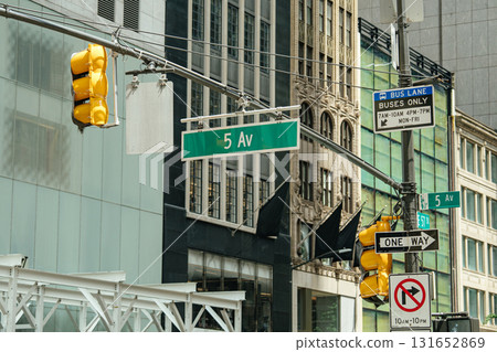 Green Fifth Avenue street signs, yellow traffic lights, and bus lane signage mark a busy New York City intersection. Iconic urban architecture frames the scene in the background. 131652869