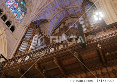 This photo showcases an elaborate pipe organ housed within a richly carved wooden balcony inside a Gothic cathedral. Above the instrument, ribbed vaulting and stained glass windows enhance the sacred 131652902