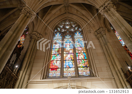 A stunning stained glass window in a gothic-style cathedral, featuring biblical scenes in vivid colors. Tall stone columns and detailed arches frame the sacred artwork, creating a majestic and 131652913