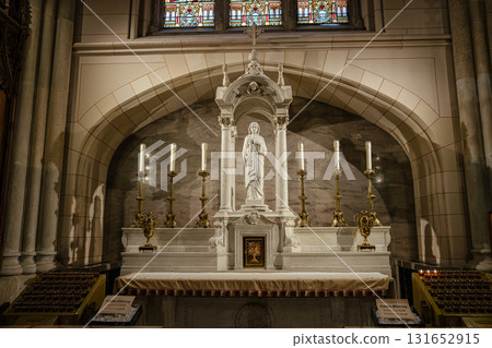 A serene marble altar featuring a central statue of Jesus Christ surrounded by tall golden candle holders. The background includes stone arches and stained glass, creating a sacred and peaceful 131652915