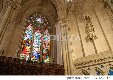 This image showcases a colorful stained-glass window depicting biblical scenes, set within a Gothic arched frame. To the right, a stone statue of a saint is mounted on the cathedral's intricately 131652916