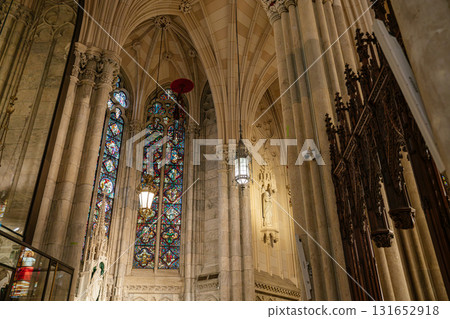 This photo captures a majestic corner of a cathedral with a towering stained-glass window framed by marble columns and vaulted arches. Ornate ironwork, a wall-mounted statue, and soft lighting add to 131652918