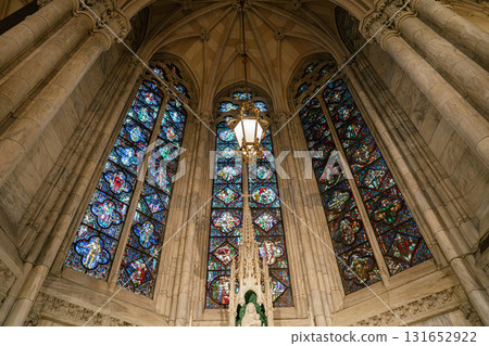 The image features a vibrant stained glass dome above a marble side altar in St. Patrick's Cathedral, New York City. Tall columns, Gothic tracery, and a golden hanging lantern add depth and richness 131652922