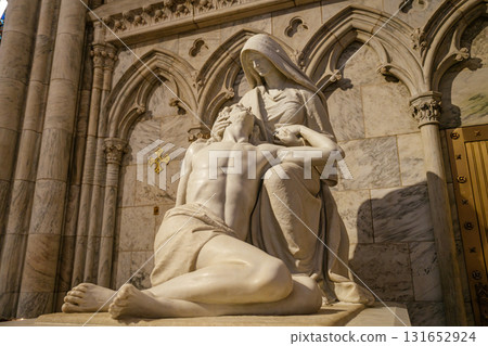 This photo captures a marble Pieta sculpture of the Virgin Mary cradling the body of Jesus, located at the entrance of St. Patrick's Cathedral in New York City. The detailed carving is set against a 131652924