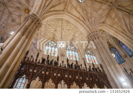 The photo highlights the ribbed vaulted ceiling, detailed choir screen, and stained glass windows inside St. Patrick's Cathedral in New York City. Ornate columns and warm lighting enhance the beauty The photo highlights the ribbed vaulted ceiling, detailed choir screen, and stained glass windows inside St. Patrick's Cathedral in New York City. Ornate columns and warm lighting enhance the beauty 131652926