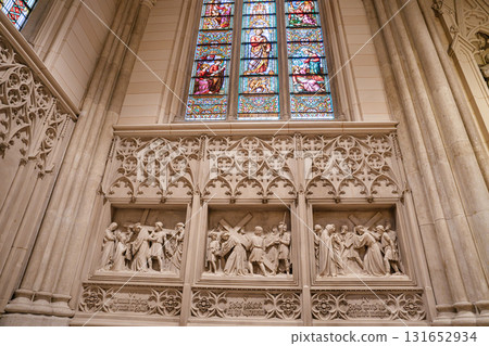 Intricate stone reliefs depicting biblical scenes are displayed beneath colorful stained glass windows inside St. Patrick's Cathedral in New York. The Gothic architecture showcases masterful 131652934