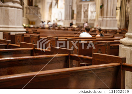 Wooden church pews fill the foreground, leading to a softly lit cathedral interior. A few people are seated, creating a quiet and reverent atmosphere in the sacred space. 131652939