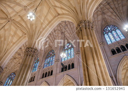 An upward view of a cathedral's vaulted ceiling showcasing intricate Gothic arches and towering columns. Vibrant stained glass windows and elegant chandeliers add beauty and light to the sacred 131652941