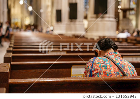 A woman in a colorful shirt is seated alone, leaning forward in silent prayer inside a cathedral. The warm wooden pews and soft lighting create a calm and spiritual atmosphere. 131652942