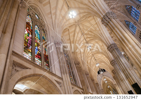 This image features intricate stained glass windows alongside soaring stone columns and arched Gothic ceilings. The warm lighting highlights the detailed architectural and artistic craftsmanship of 131652947