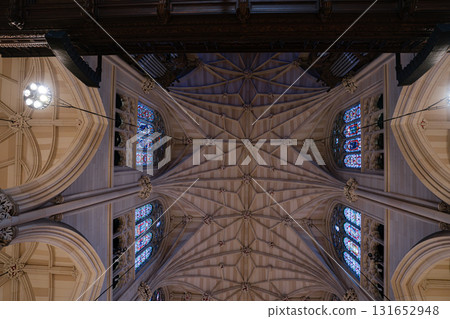 A symmetrical view of a gothic vaulted ceiling with ribbed arches and colorful stained glass windows. The stonework and stained panels reveal intricate craftsmanship inside the cathedral. A symmetrical view of a gothic vaulted ceiling with ribbed arches and colorful stained glass windows. The stonework and stained panels reveal intricate craftsmanship inside the cathedral. 131652948