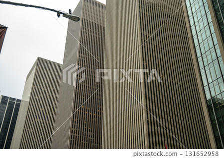 A group of tall skyscrapers with narrow vertical windows rises in Midtown Manhattan. The sharp vertical patterns create a powerful corporate atmosphere against the cloudy sky. A group of tall skyscrapers with narrow vertical windows rises in Midtown Manhattan. The sharp vertical patterns create a powerful corporate atmosphere against the cloudy sky. 131652958