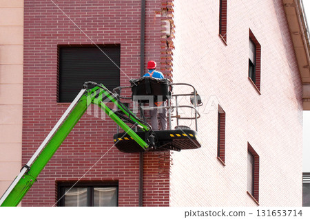 Construction worker in uniform and red cap repairing exterior brick corner wall of residential building in aerial lift basket Construction worker in uniform and red cap repairing exterior brick corner wall of residential building in aerial lift basket 131653714