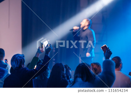 Live concert audience cheers as singer performs under blue spotlight with fans filming on phones Live concert audience cheers as singer performs under blue spotlight with fans filming on phones 131654234