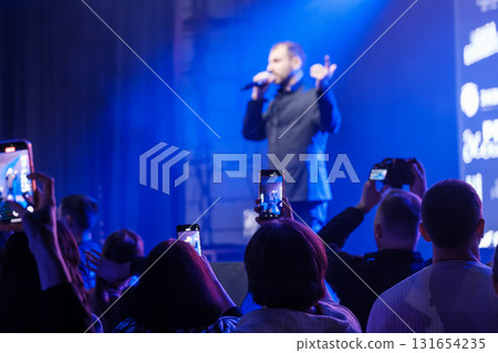 Live concert performance by a singer on stage under blue lights with crowd filming on smartphones Live concert performance by a singer on stage under blue lights with crowd filming on smartphones 131654235