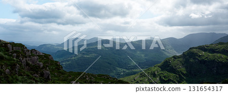 A panoramic view of rolling green hills and mountains, some capped with rocks, in the Gap of Dunloe in County Kerry, Ireland, under a cloudy sky during the daytime. A panoramic view of rolling green hills and mountains, some capped with rocks, in the Gap of Dunloe in County Kerry, Ireland, under a cloudy sky during the daytime. 131654317