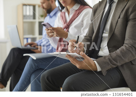 Business man writing on clipboard while others are using laptops and phones in background 131654401