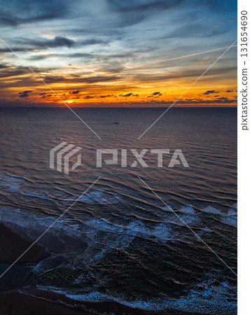 Aerial view of coastal sunset with golden light reflecting on gentle waves shoreline foam and distant vessel add depth to the serene ocean horizon beneath textured clouds. Aerial view of coastal sunset with golden light reflecting on gentle waves shoreline foam and distant vessel add depth to the serene ocean horizon beneath textured clouds. 131654690