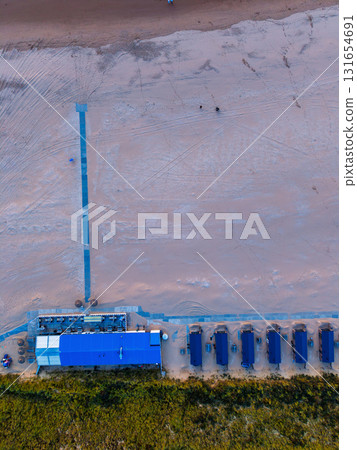 Aerial view of beachside facility with blue roofed cabins and main building boardwalk cuts across sandy shore toward calm sea, highlighting geometric layout and coastal infrastructure. Aerial view of beachside facility with blue roofed cabins and main building boardwalk cuts across sandy shore toward calm sea, highlighting geometric layout and coastal infrastructure. 131654691