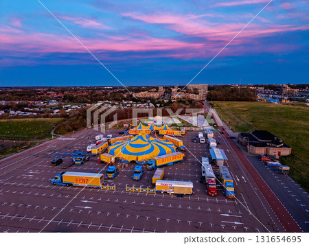 Aerial view of vibrant circus setup in sunset lit parking lot yellow blue tent, trailers, and trucks contrast with pink sky and forested backdrop, blending spectacle with serene surroundings. 131654695