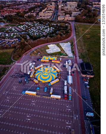 Aerial view of circus setup in parking lot beside skate park and residential zone yellow blue tent, trailers, and ramps contrast with muted urban tones and green spaces. 131654696