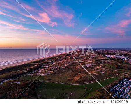 Aerial view of coastal landscape at sunrise pink and purple sky over ocean, sandy beach, grassy dunes, and nearby town highlight contrast between natural beauty and urban development. Aerial view of coastal landscape at sunrise pink and purple sky over ocean, sandy beach, grassy dunes, and nearby town highlight contrast between natural beauty and urban development. 131654699