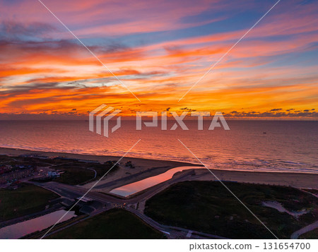 Aerial view of coastal sunset with vibrant sky over calm sea river inlet flows into ocean beside sandy beach and scattered buildings, blending warm light with tranquil natural geometry. 131654700