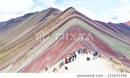 Tourists hike and gather at the summit of Peruan Rainbow Mountain, admiring its colored mineral striped slopes under a bright sky, with a sweeping view of the unique geological formation. 131655398