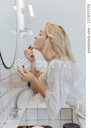 Blonde Young Woman Applying Nude Lipstick On Lips While Standing In Front Of Mirror In Bathroom 131655559