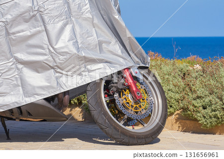 Detail of brake disc and tire peeking from silver tarp on parked motorbike near blue sea under bright sky 131656961