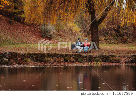 Son and older father sitting on bench by lake and talking. 131657599
