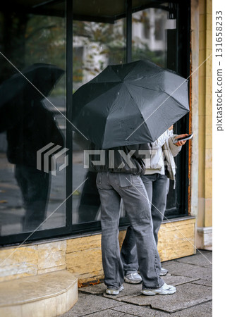 Two people standing under black umbrella on rainy city street, soft selective focus. Autumn City Mood. Concept of urban lifestyle, rainy mood, wet weather 131658233