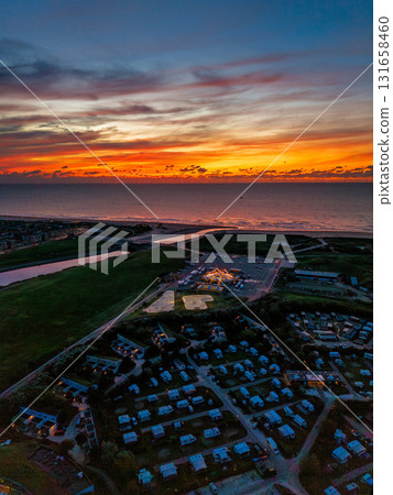 Aerial view of coastal town at sunset grid of houses and trailers, bright central hub, and canal flowing into ocean beneath vivid orange blue sky, blending structure with natural drama. Aerial view of coastal town at sunset grid of houses and trailers, bright central hub, and canal flowing into ocean beneath vivid orange blue sky, blending structure with natural drama. 131658460