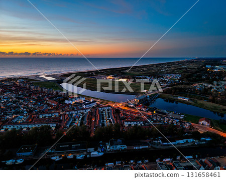Aerial view of coastal town at sunset dense housing grid, illuminated roundabout, canal flowing to ocean, and vibrant sky over dunes blend urban order with natural serenity. Aerial view of coastal town at sunset dense housing grid, illuminated roundabout, canal flowing to ocean, and vibrant sky over dunes blend urban order with natural serenity. 131658461