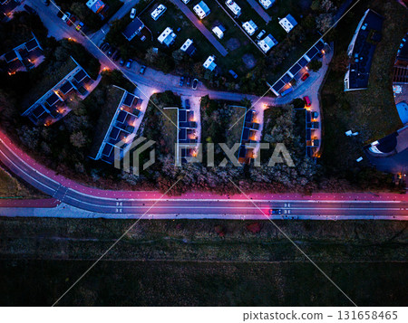 Aerial view of twilight residential zone curved housing grid, lit multi lane road with red bike paths, and grassy field below blend geometric planning with natural contrast. 131658465
