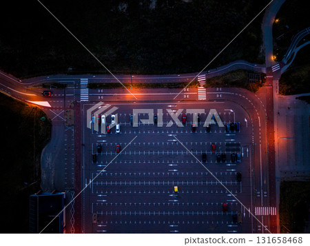 Aerial nighttime view of empty parking lot warm orange streetlights illuminate marked lanes and scattered cars, highlighting symmetry and contrast with surrounding darkness. 131658468