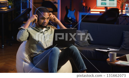 Black person listening to focus music on headphones to finish tasks, multitasking in his living room on the bean bag. Young adult enjoying calm study sounds to complete assignments. Camera b. 131662073