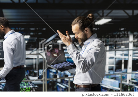 Data center male developer frustrated by critical error affecting laptop, worrying about security breach and criminal activity. Worker reacting to hardware performance issues triggered by bugs. Data center male developer frustrated by critical error affecting laptop, worrying about security breach and criminal activity. Worker reacting to hardware performance issues triggered by bugs. 131662365